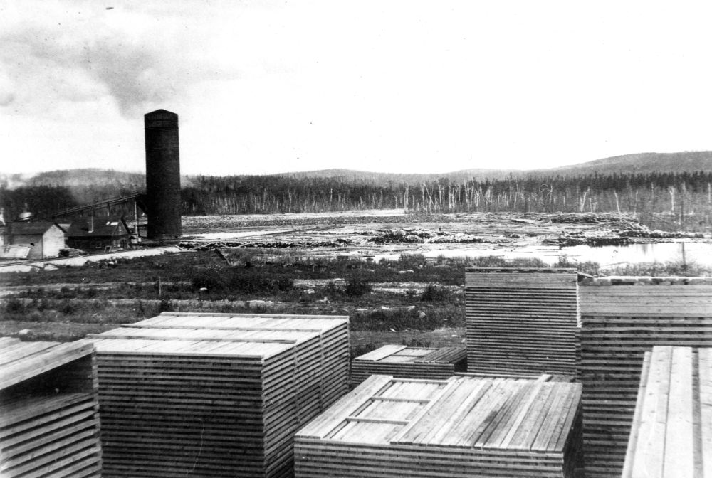 Black and white photograph of a wood yard: numerous neatly piled stacks of wooden planks. Background: factory, tall chimney, smoke, expanse of water, numerous floating logs. Scene framed by trees and distant hills.