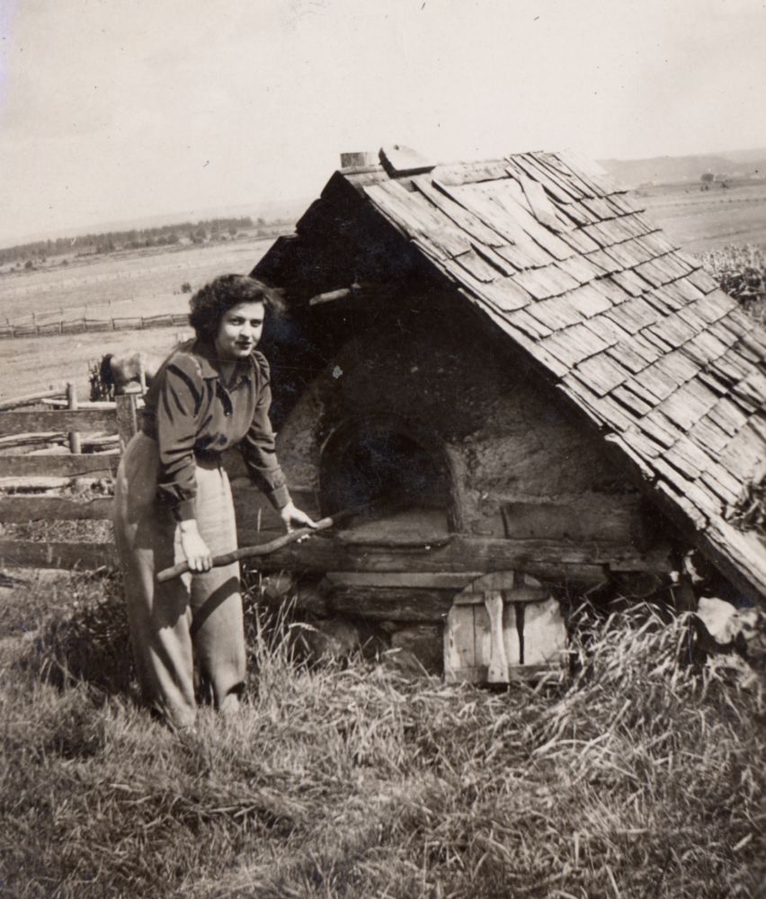 Black and white photograph: woman standing near an outdoor bread oven. She holds a long wooden stick. Oven made of wood and stone, worn shingle roof. Fence in the background, distant rural landscape. Atmosphere of a time when outdoor cooking was common.