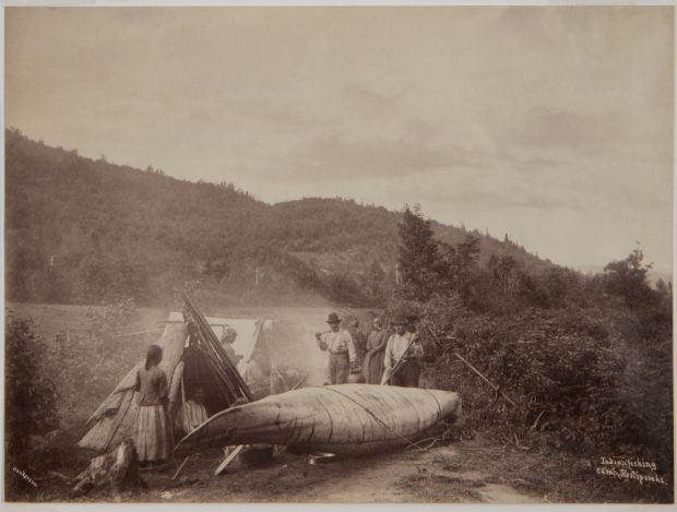 Albumen print (around 1870): Indigenous fishing camp, Restigouche River, QC-NB. Campsite by the river (QC-NB border). Foreground: birch bark canoe near bushes. Left: conical structure (wigwam/traditional shelter), several people. Centre: fire (thick smoke). Background: wooded hill under a cloudy sky. Bottom right: “Indian fishing camp, Restigouche.”