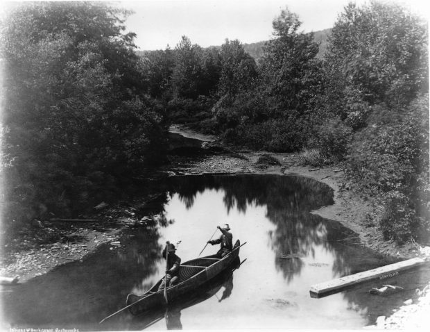 Albumen print (around 1870, Alexander Henderson): Two Indigenous Men in a Bark Canoe, Restigouche, QC (peaceful winding river scene). Two individuals navigate in a birch bark canoe. Dark water mirror effect. Lush vegetation (conifers, hardwoods).