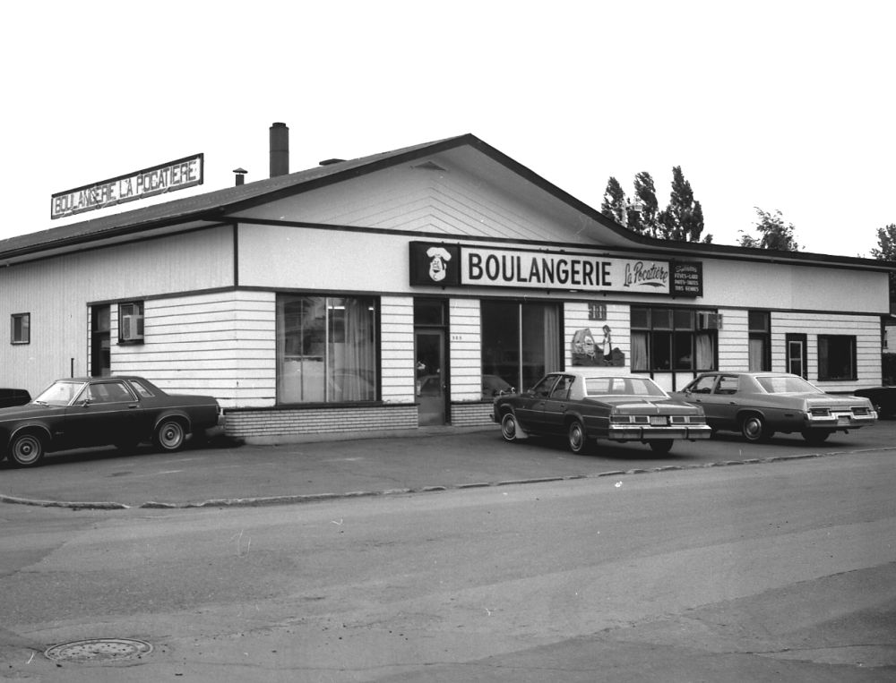 Black and white photograph: facade of a commercial building "BOULANGERIE & PÂTISSERIE" (Bakery & Pastry Shop). Single-story building, light horizontal siding, flat roof with a slight slope. Large storefront windows. Several cars parked in front (paved). Photo from the early 1970s.