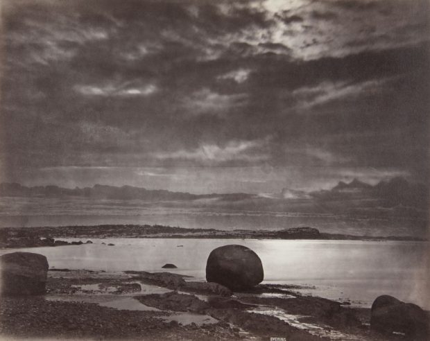 Albumen print (1876, Alexander Henderson): Evening on the beach at Métis (melancholy twilight seascape). Dominant sky (dark textured clouds, openings of light). Foreground: wet rocky beach (large dark rocks including a rounded block in the centre right).