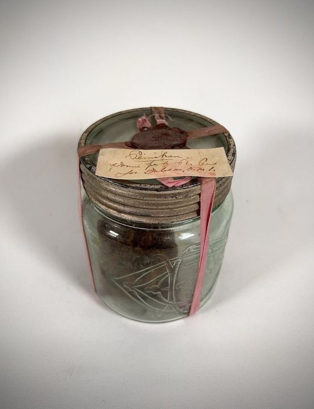 Clear cylindrical glass jar filled with pemmican. Screw-on metal lid. Yellowed paper (handwritten note in brown ink) attached to the lid with a faded pink ribbon and red wax seal.