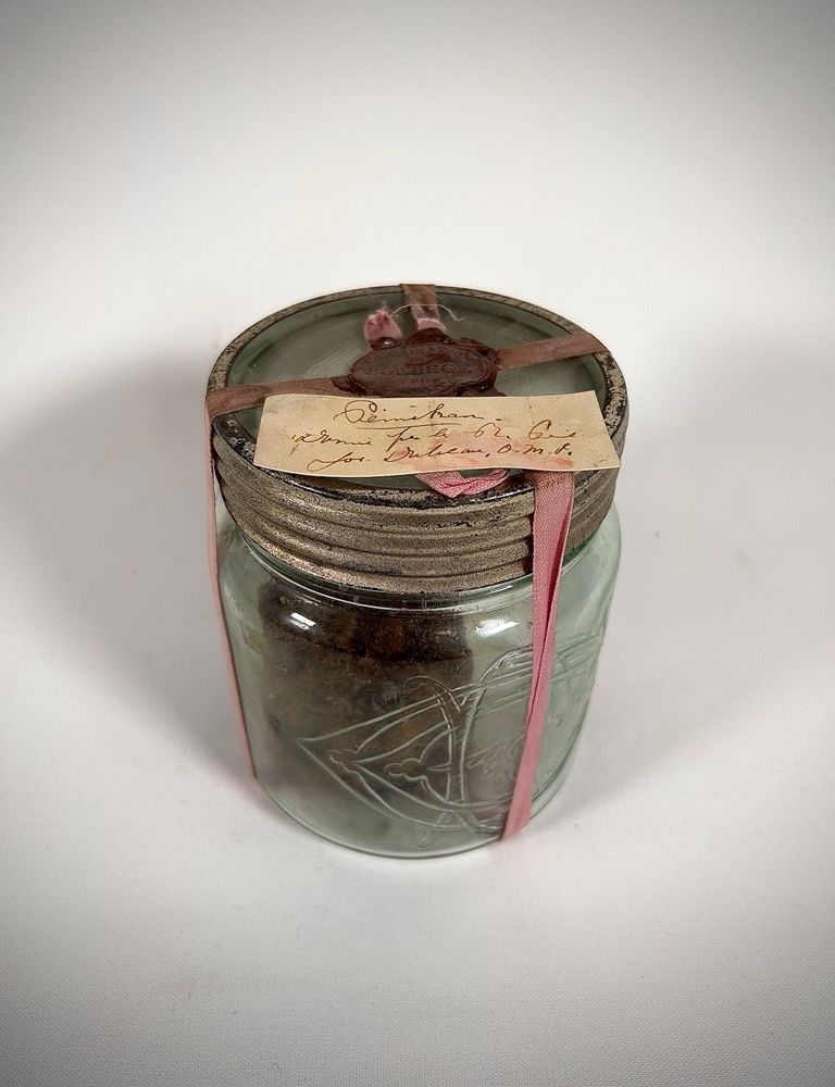 Clear cylindrical glass jar filled with pemmican. Screw-on metal lid. Yellowed paper (handwritten note in brown ink) attached to the lid with a faded pink ribbon and red wax seal.