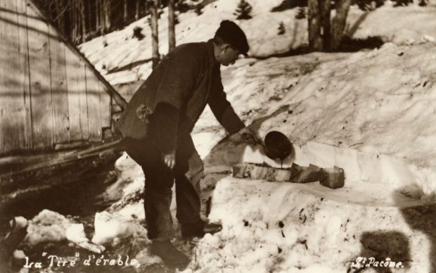 Sepia photograph, making maple taffy (S. Pacôme): a man in winter clothing pours maple taffy into bark containers on the snow. Background: snow-covered woods, part of a wooden structure on the left.