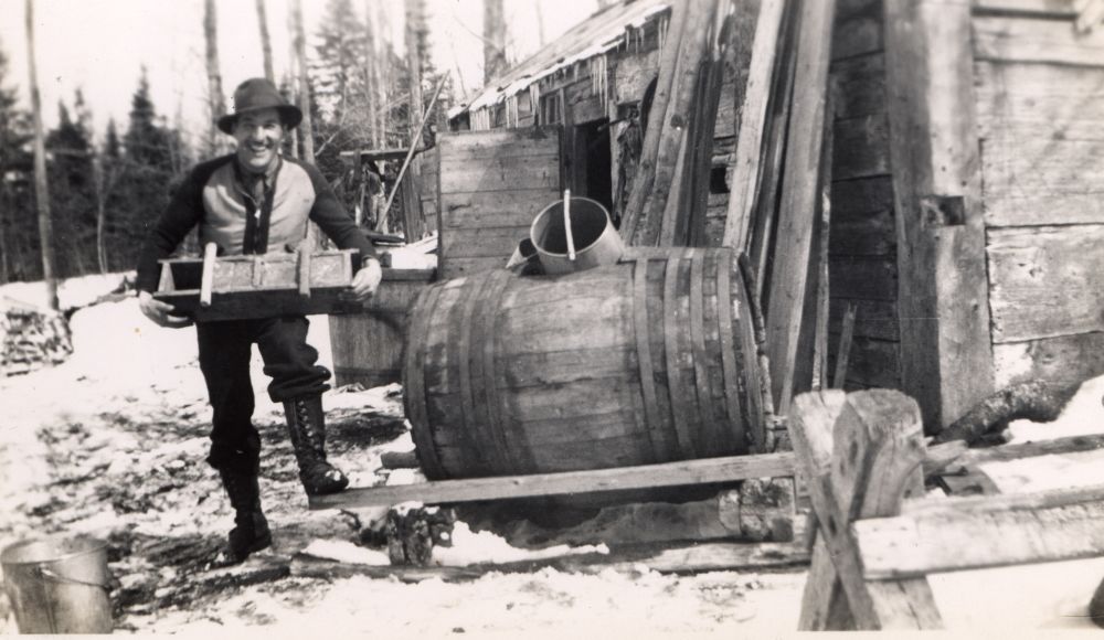 Sepia photograph: smiling man carrying sugar moulds. Snowy environment, near a rustic wooden sugar shack and a large wooden barrel on supports.