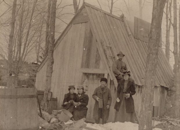 Sepia photograph: family celebration in front of a rustic wood cabin (wooded and snowy environment). Left foreground: two young women sitting. Centre: young man standing. Right: woman standing. Cabin roof: seated man . Far left: large wooden barrel.
