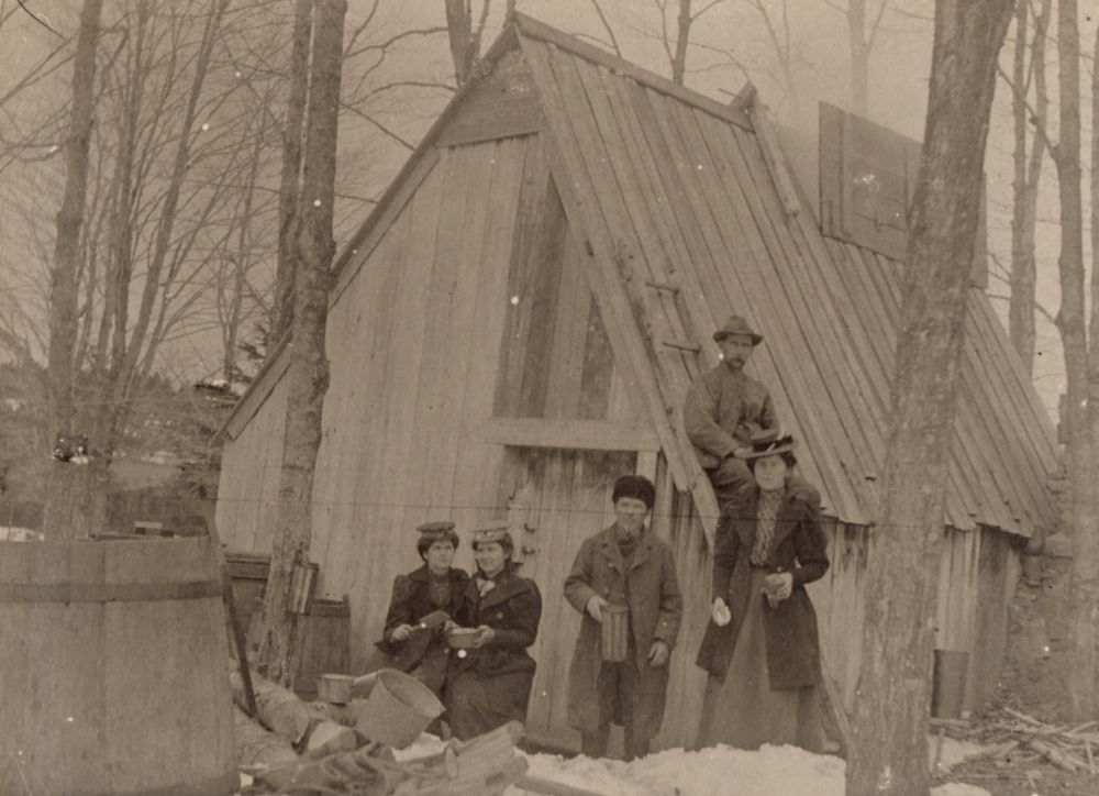 Sepia photograph: family celebration in front of a rustic wood cabin (wooded and snowy environment). Left foreground: two young women sitting. Centre: young man standing. Right: woman standing. Cabin roof: seated man . Far left: large wooden barrel.