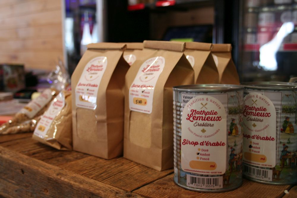 Brown paper bags, maple nuggets; two cans of syrup from "Nathalie Lemieux Érablière". Foreground: cans with labels depicting maple harvesting scenes.
