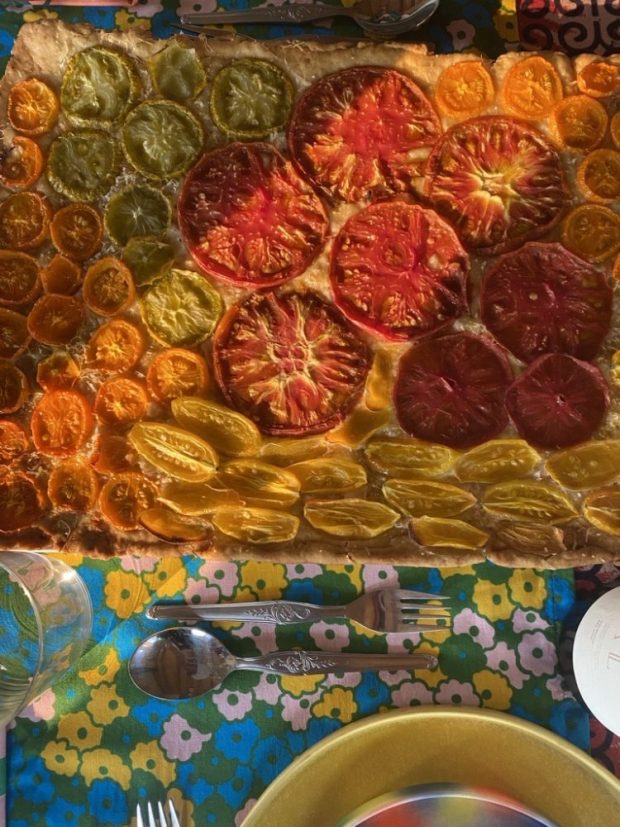Rectangular savory tart or focaccia topped with multi-colored tomato slices (red, orange, yellow, green) placed on a colorful floral-patterned tablecloth.