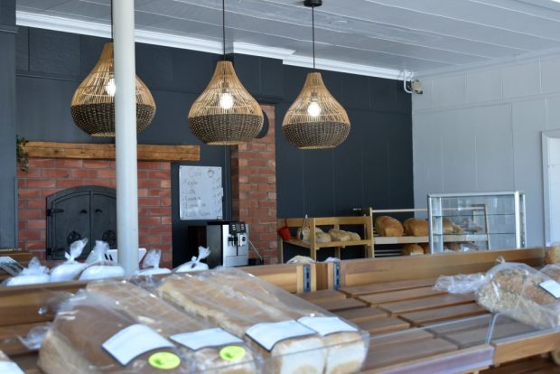 Colour photograph: Interior of a traditional bakery. In the foreground, packaged bread is displayed on a wooden rack. In the background, a brick oven and hanging wicker light fixtures contribute to the bakery’s rustic and authentic atmosphere.