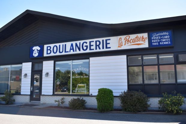 Colour photograph: Façade of the Boulangerie La Pocatière. The bright sign, bearing a chef’s logo, lists specialties offered, such as baked beans and pies. The dark building is adorned with white siding and large windows.