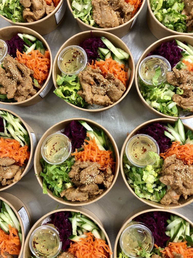 Overhead shot of a series of individual cardboard bowls filled with crispy chicken rice vermicelli. The bowls are generously topped with lettuce, shredded carrots, julienned purple vegetables (cabbage or beetroot), and contain a small pot of dressing.