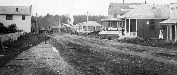 A black and white photograph of a wide dirt road with buildings on both sides of it. There is a person standing outside the entrance of two buildings on the right and a group of people visible down the road near an old automobile.