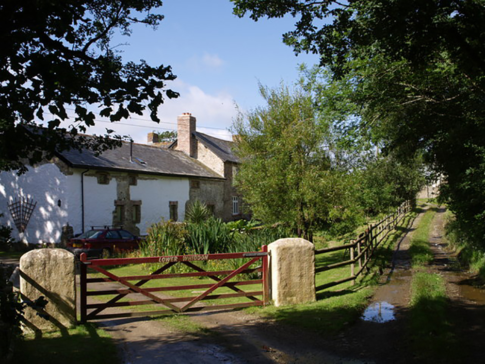 A rural gravel road runs alongside the Bickles farmhouse. The road is lined by a wooden fence that transitions to a large red gate in front of the country home. The gate has white text on it that reads, “Lower Whiddon.”