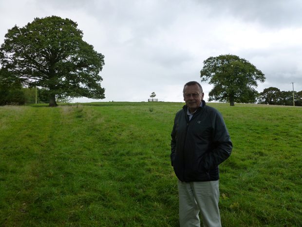 Tom Pope stands in a grass field that has an impression of an old trail in the grass. There are two large trees and a wooden fence visible in the background.