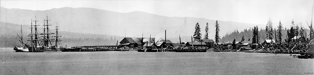 A black and white photograph of the Hastings Sawmill in 1872. The mill is located along the shoreline and has a number of small buildings around it. There is a European ship docked at the port on the left side of the image.