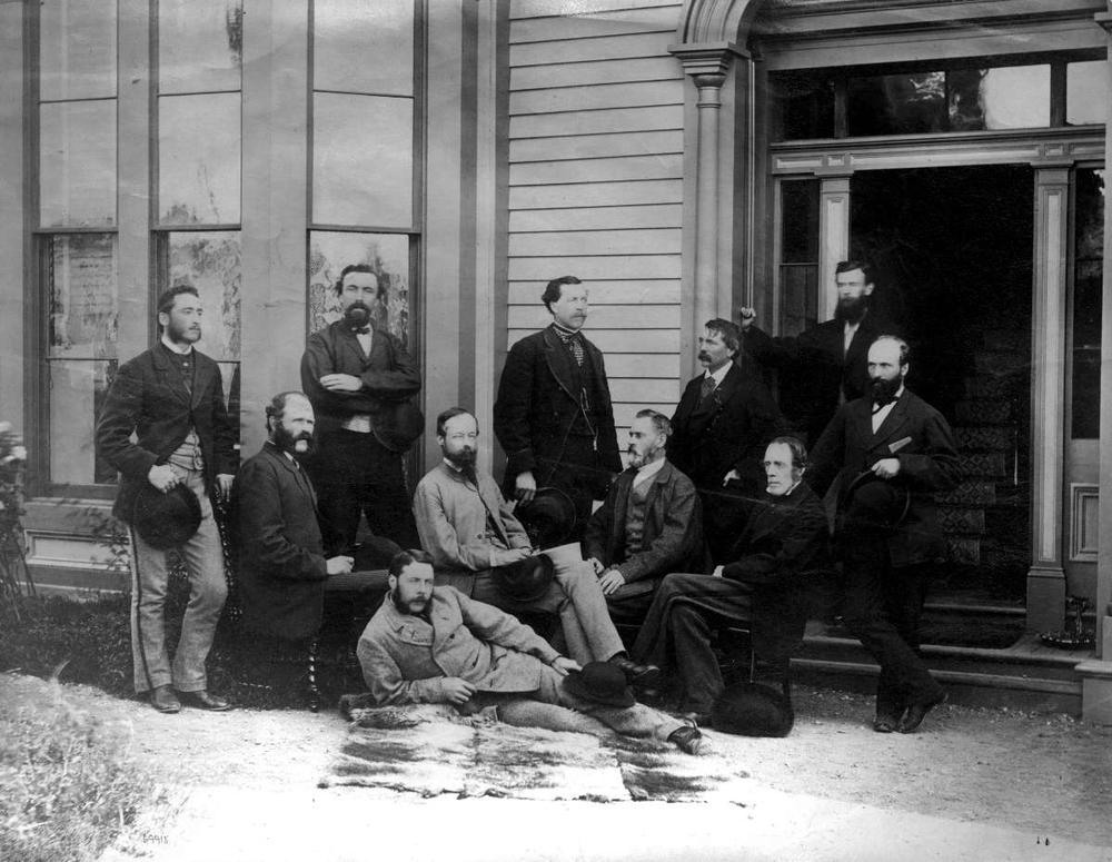 A black and white photograph of Walter Moberly and ten other men of a CPR Survey Party. The men are wearing suits and are organized into three approximate rows in front of a building. One gentlemen is laying on his side in the front row, the second row consists of four seated men, and the third consists of six men standing.