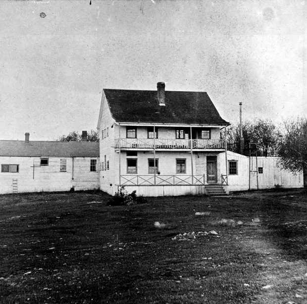 A black and white photograph of the Victoria Lunatic Asylum. The main building is two storeys with a porch on the first level and a deck on second. There is a long one story building directly behind the front building.