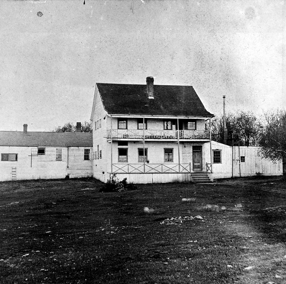 A black and white photograph of the Victoria Lunatic Asylum. The main building is two storeys with a porch on the first level and a deck on second. There is a long one story building directly behind the front building.