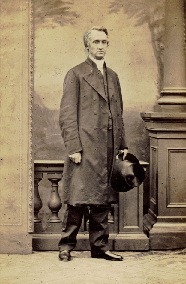 A black and white studio portrait photograph of a Bishop George Hills wearing a long coat. He is holding a top hat in his left hand and has his right hand clenched at his side.