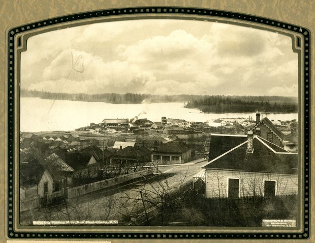 A sepia toned photograph taken from a high perspective of New Westminster that shows a number of houses and the Royal City Planing Mills along the Fraser River.