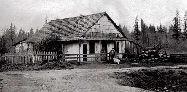 A black and white photograph of the Jackman’s first Aldergrove house. The house is light coloured with a wooden shingle roof, a picket fence on the left side, and a wooden fence in front of the house. Jackman and a light coloured dog are standing in front of the house.
