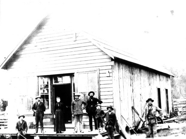 A black and white photograph of six men, one woman, and one boy standing in front of Shortreed’s Store. The store is a large wooden building with a front patio that leads to the front door that has a window on each side of it. There are two additional doors visible along the back right side of the building.