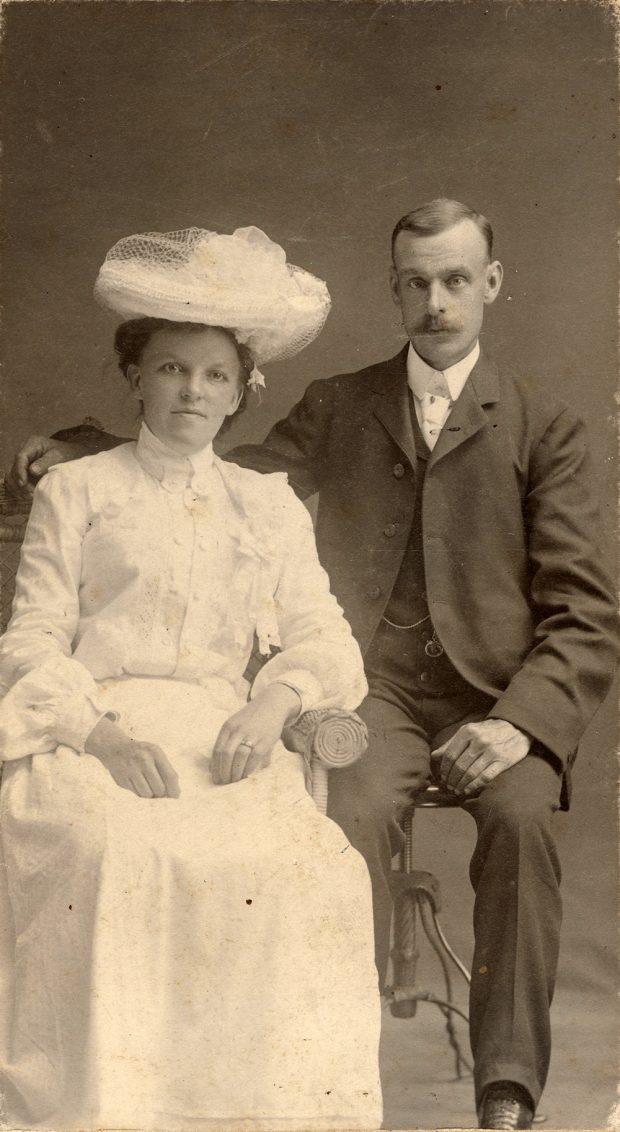 A sepia toned studio portrait photograph of Philip Jackman Junior and Anna Caroline Jackman (née Wadel). The two are seated on separate chairs and Philip has his right arm behind Anna, on the back of her chair. Anna is wearing a white dress and hat and Philip Jr. is wearing a suit.