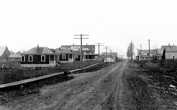 A black and white photograph of downtown Aldergrove circa 1900. A dirt road runs through the small town that has buildings on both sides of the road.