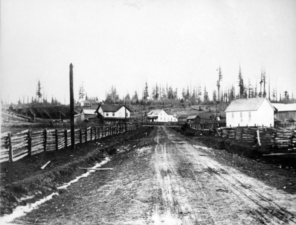 A black and white photograph of Murray’s Corners from the Old Yale Road in Langley. The road goes up a hill and is lined with wooden fences on both sides. There are four large buildings at the top of the hill.
