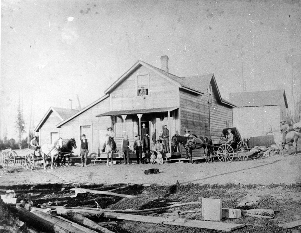 Photographie en noir et blanc d’un groupe de personnes devant le magasin de Davidson et Riddell à Murray’s Corner, à Langley. C’est un grand bâtiment à deux étages, en bois. Le groupe est constitué de deux voitures tirées par des chevaux, un homme à cheval, et dix autres personnes assises ou debout devant le bâtiment.