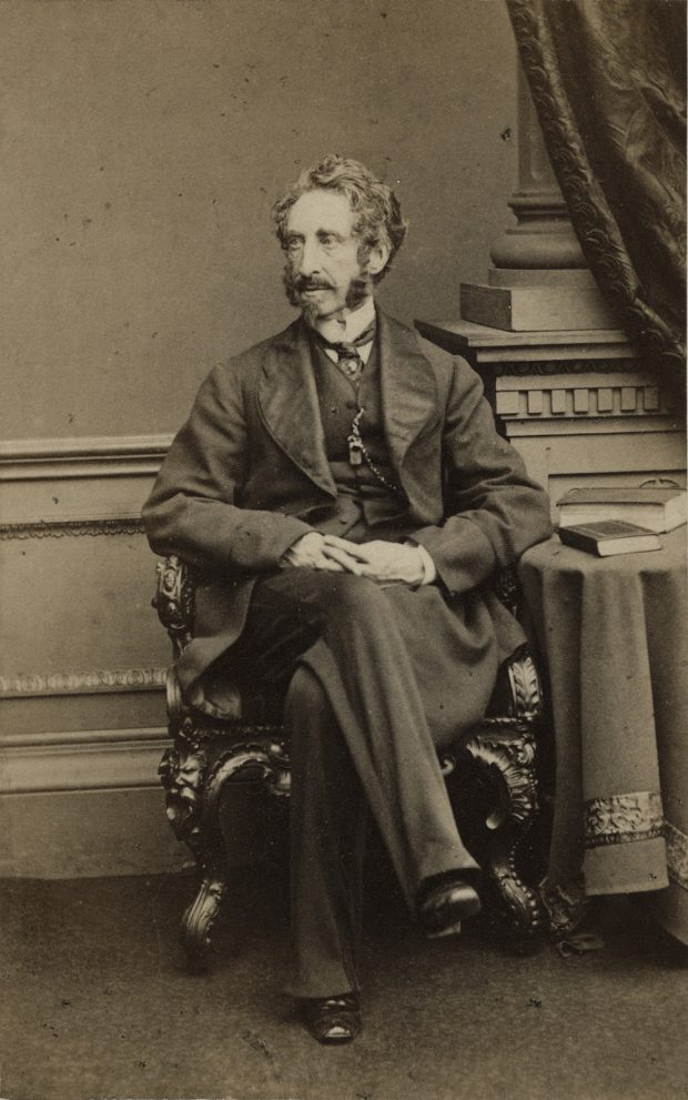A black and white studio portrait of Sir Edward Bulwer-Lytton sitting on a decorative chair next to a side table with two books on it.