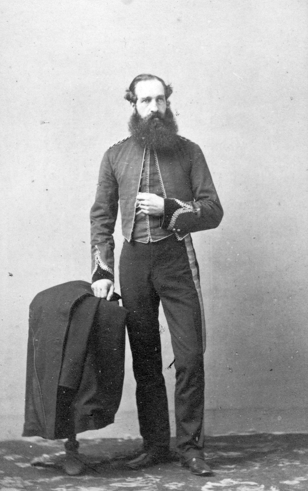 A black and white studio portrait of Captain Henry Reynolds Luard with a large beard. Luard is wearing a military uniform and has his left arm bent across the front of his body, and his right hand on his coat, which is laying on top of a small table.