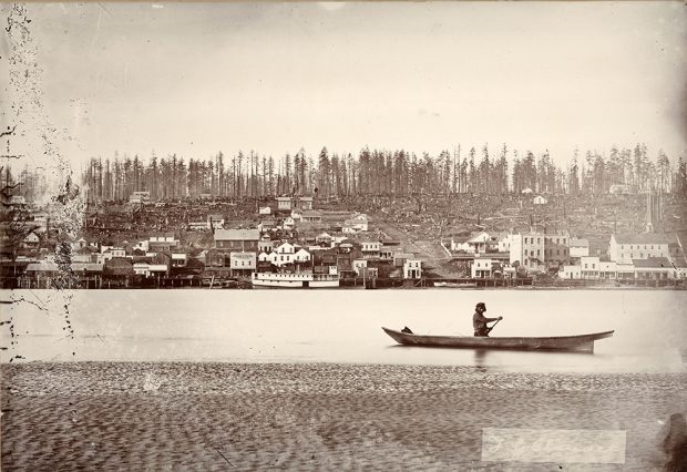 A sepia toned photograph of a man canoeing on the Fraser River in front of a recently deforested New Westminster. New Westminster has multiple buildings along the river, but the buildings become scarcer further up the hill. The upper portion of the hill is littered with tree stumps.