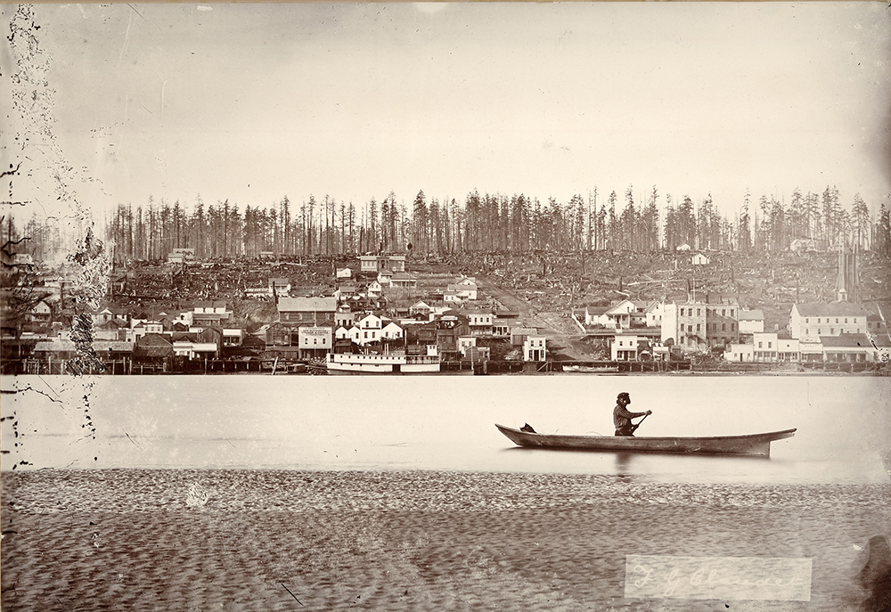 Une photographie monochrome aux tons sépia d’un homme en canot sur le fleuve Fraser passant devant une partie récemment déboisée de New Westminster. New Westminster, à cette époque, compte de nombreux bâtiments près de la rive du fleuve mais, plus on s’éloigne de la rive, moins il y a d’édifices. Le haut des collines est couvert de souches d’arbres