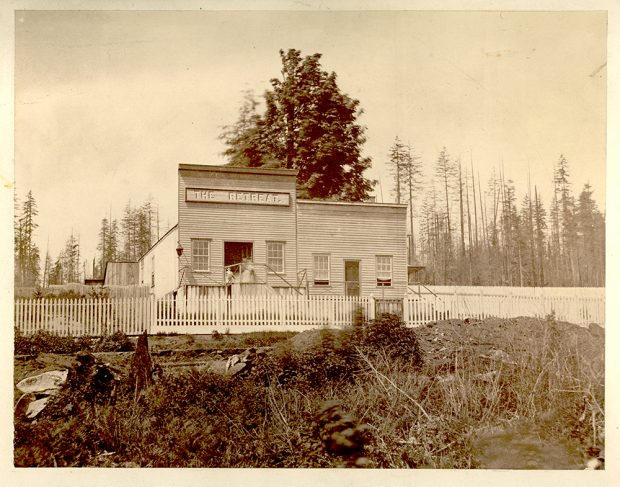 A sepia toned photograph taken circa 1865 of a building with two storefronts. The storefront on the left is a saloon which has a taller false front with a sign on it that reads, The Retreat. Each storefront has a door in the centre with a window on each side of the door. There is a picket fence in front of the building and a forest in the background.