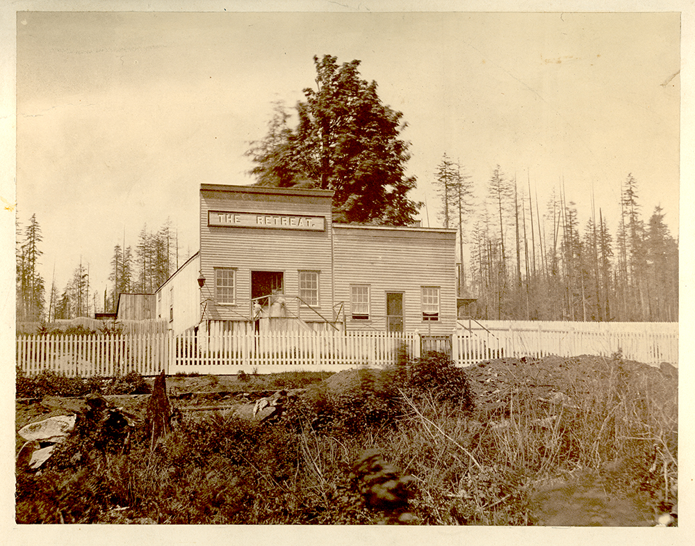 A sepia toned photograph taken circa 1865 of a building with two storefronts. The storefront on the left is a saloon which has a taller false front with a sign on it that reads, "The Retreat." Each storefront has a door in the centre with a window on each side of the door. There is a picket fence in front of the building and a forest in the background.