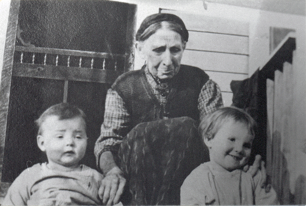 Une photographie en noir et blanc, prise en 1917, de Sarah Ann Jackman, âgée, sur le perron avec ses deux petits-enfants. Sarah a les mains sur les épaules des enfants.