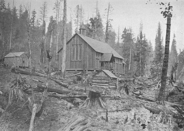 A black and white photograph of the Trinity Church and two other small wooden buildings around it. The buildings are situated in a recently logged plot of land with felled trees and stumps littered throughout the foreground