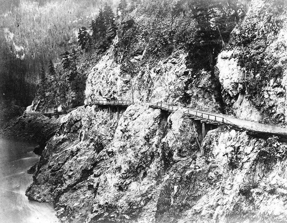 A black and white photograph of a narrow wagon road built along a rocky cliff that sits above a river. Sections of the road are made up of bridges with wooden railings on one side.