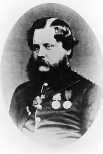 A black and white oval shaped bust portrait of Dr. John Vernon Seddall, dressed in a formal military outfit with an assortment of medals and achievements across his chest. Seddall has slick parted hair and a large beard.