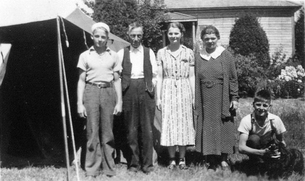 A black and white photograph of a group of two girls, two boys, and an elderly man beside a tent in front of a house. All are standing except for one boy on the right who is kneeling and petting a dog.