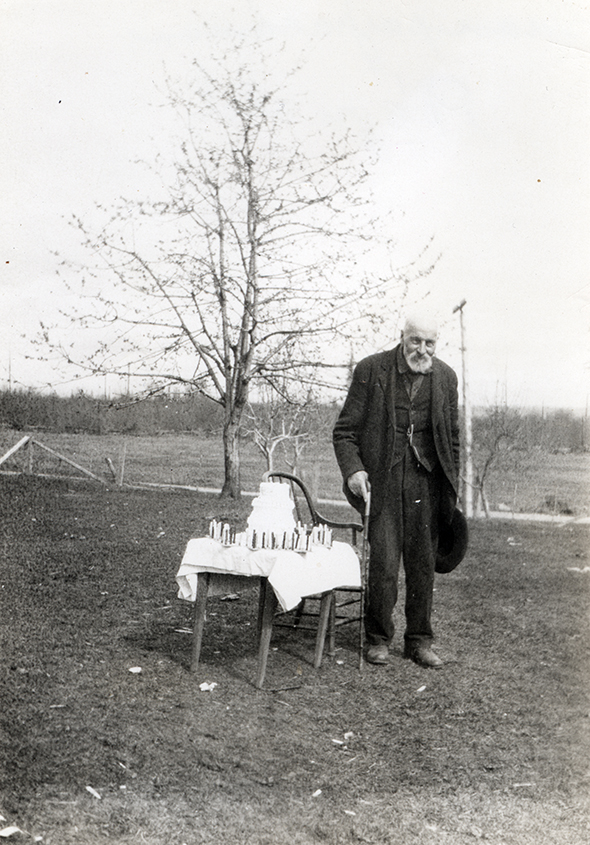 A sepia toned photograph of an elderly man standing beside a wooden table with a three tiered cake on it.