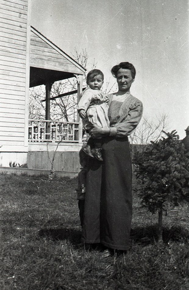 A black and white photograph of a woman holding a small boy in her arms standing in the grass in front of a house.