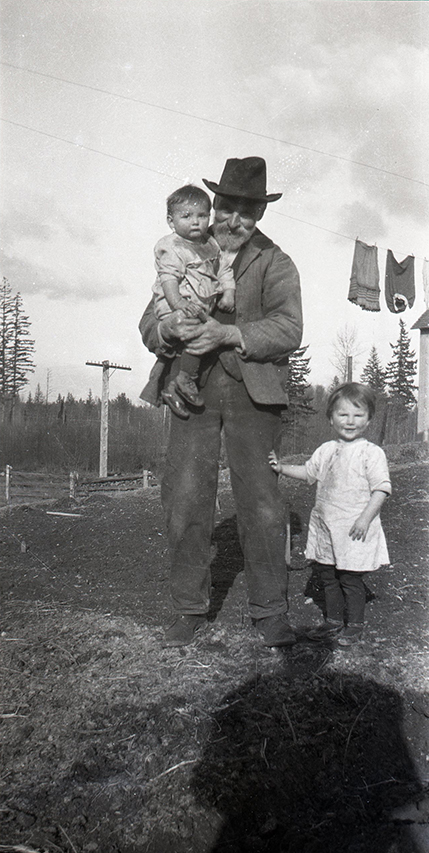 A black and white photograph of a man holding a small boy in his arms and a small girl standing beside him. The small girl has on hand on the man’s leg. There is a clothing line strung above the people with a few articles of clothing hanging from it on the right side.