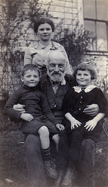 A sepia toned photograph of a man sitting on a chair with a boy and a girl sitting on his lap. There is a woman standing behind them with a building visible in the background.
