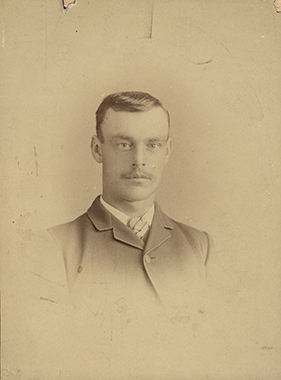 A sepia toned bust studio portrait photograph of a young man with a moustache.
