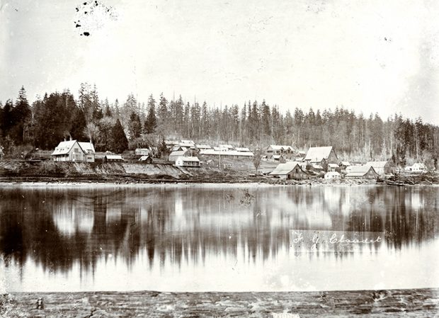 A black and white photograph of a small settlement along a river. The settlement consist of a grouping of small buildings that is surrounded by thick forest beyond it.