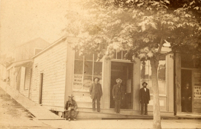 Une photographie de quatre hommes, trois debout et un assis, devant le « Grotto Saloon » à New Westminster. Une enseigne visible dans l’une des fenêtres porte les mots : « Bière Vin Spiritueux ». 